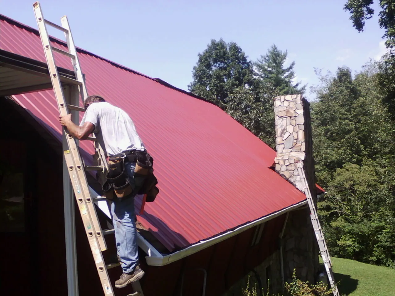 Roofer on a ladder beside a red metal roof and stone chimney — Soffit & Fascia Repair by Roofing Brothers in Wheatcroft